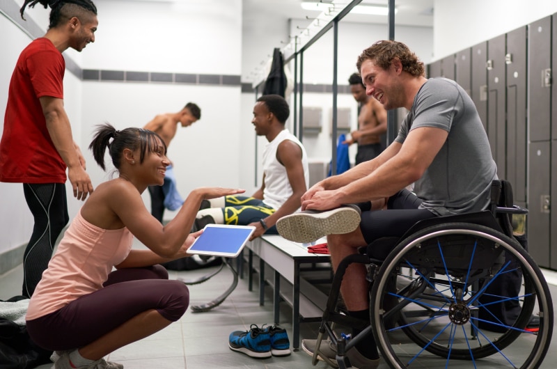 Athletes relax and laugh together in a locker room, including a man in a wheelchair and a woman holding a tablet.