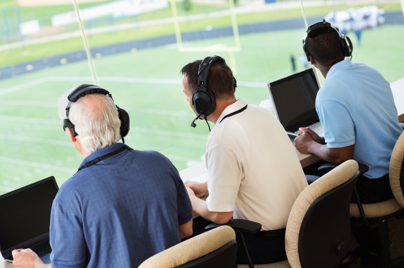 Three sports announcers in headsets watch and commentating from a press box overlooking a football field.