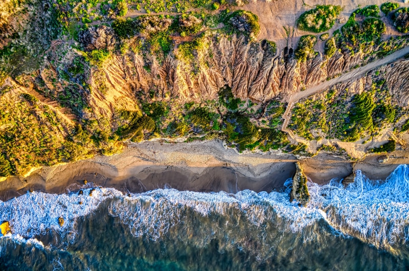 Aerial view of a California coastline where ocean waves meet cliffs and green hills under golden sunlight.
