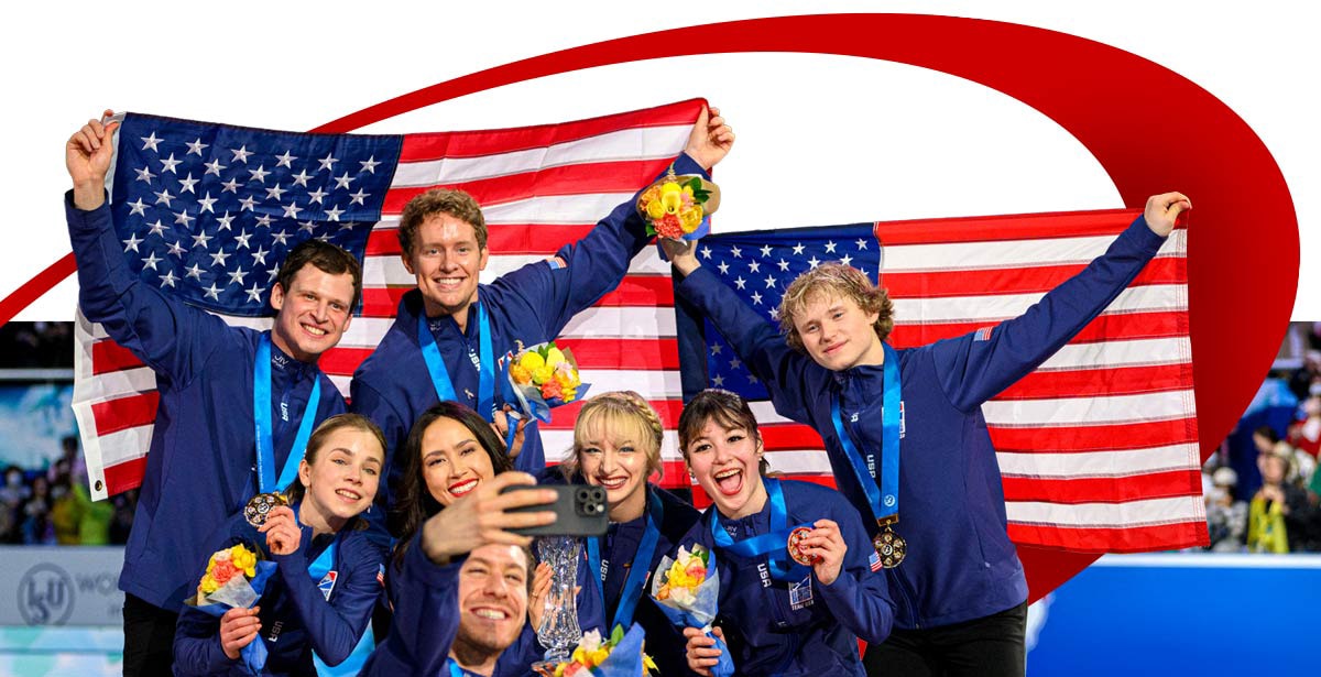 Team USA skaters celebrate with medals and bouquets, holding American flags and taking a selfie at an ice arena.