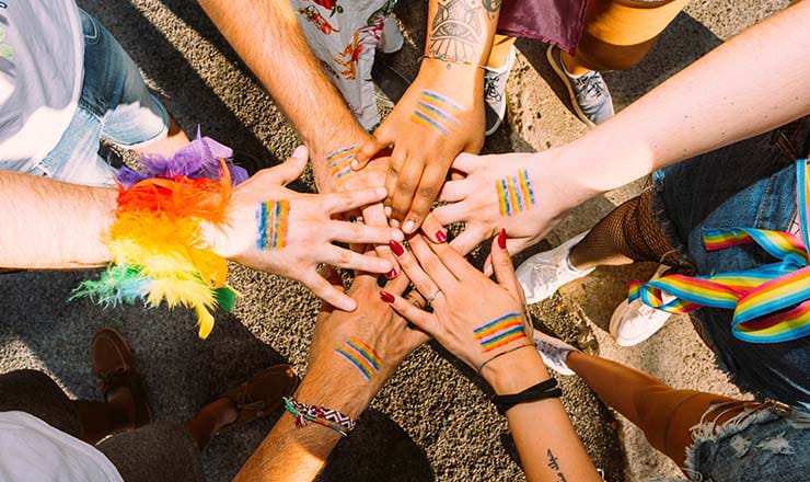ix hands decorated with LGBTQ+ Pride flag stacked on top of each other with wrists decorated in rainbow feathers and ribbons
