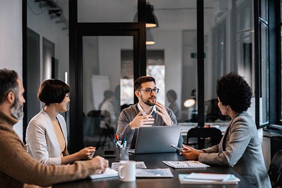Four colleagues discuss information around a laptop in a glass-walled conference room