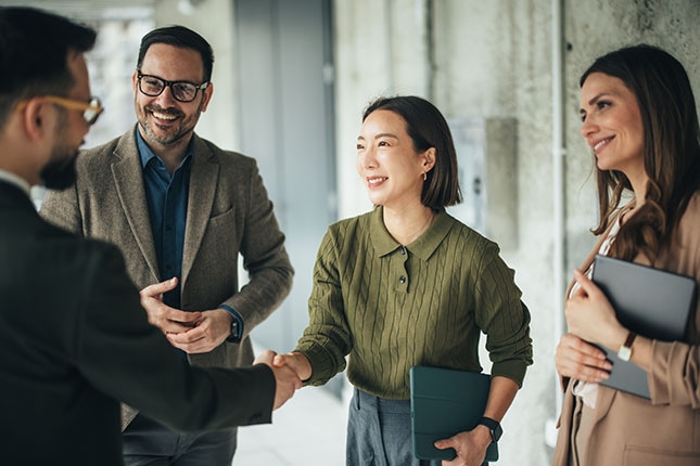 Four professionals meet in an office setting, shaking hands and holding notebooks and tablets during a collaborative discussion.