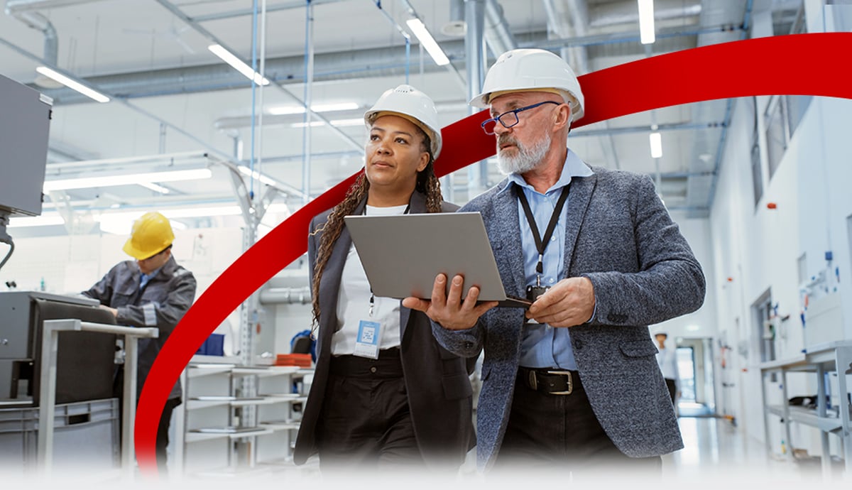 Two professional manufacturing engineers wearing hardhats carrying a laptop.