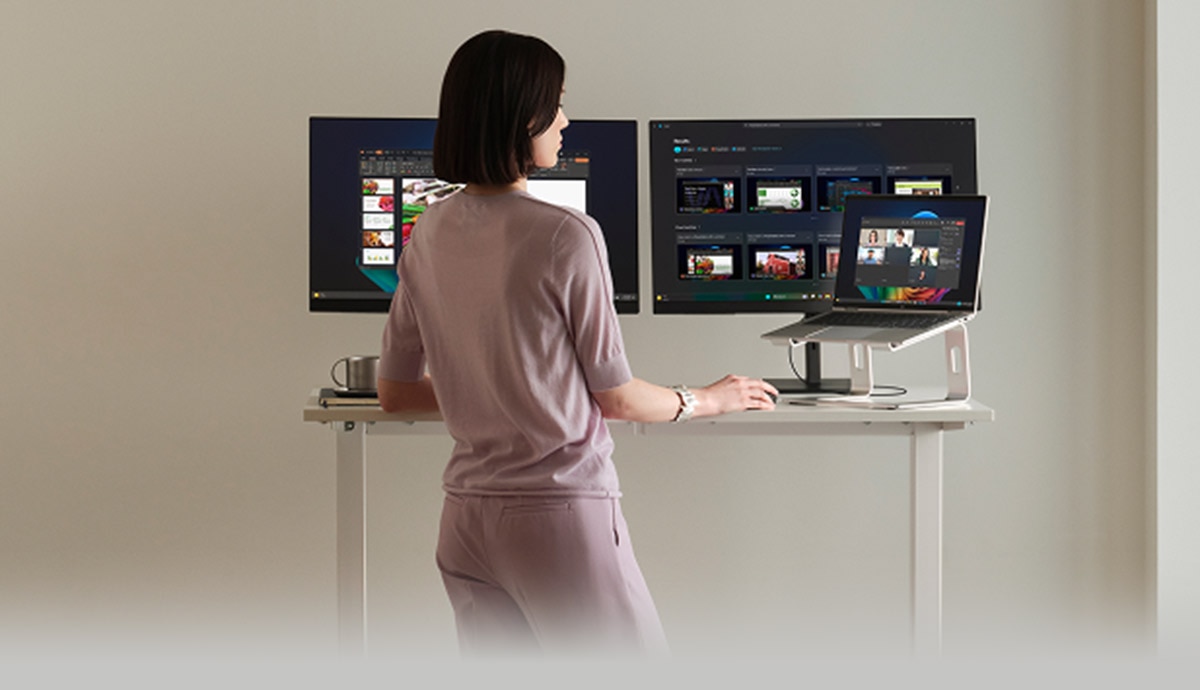 Modern workspace with a person standing at an adjustable desk using a Copilot+ PC connected to two external monitors, displaying multiple applications and illustrating AI-powered workflows, performance, and multitasking on Windows 11