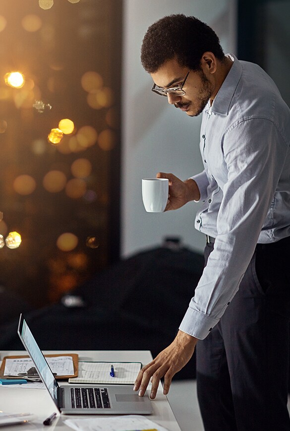 Image of a man standing at a desk holding a cup and using a laptop.
