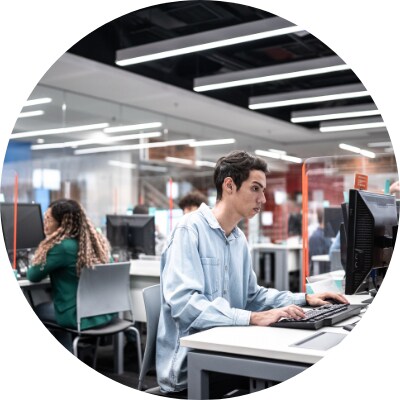 Young professional working on a computer at his office work station with other colleagues in the background.