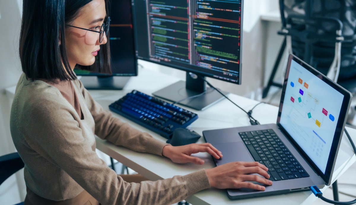 A female IT professional works at a desk surrounded by multiple monitors.