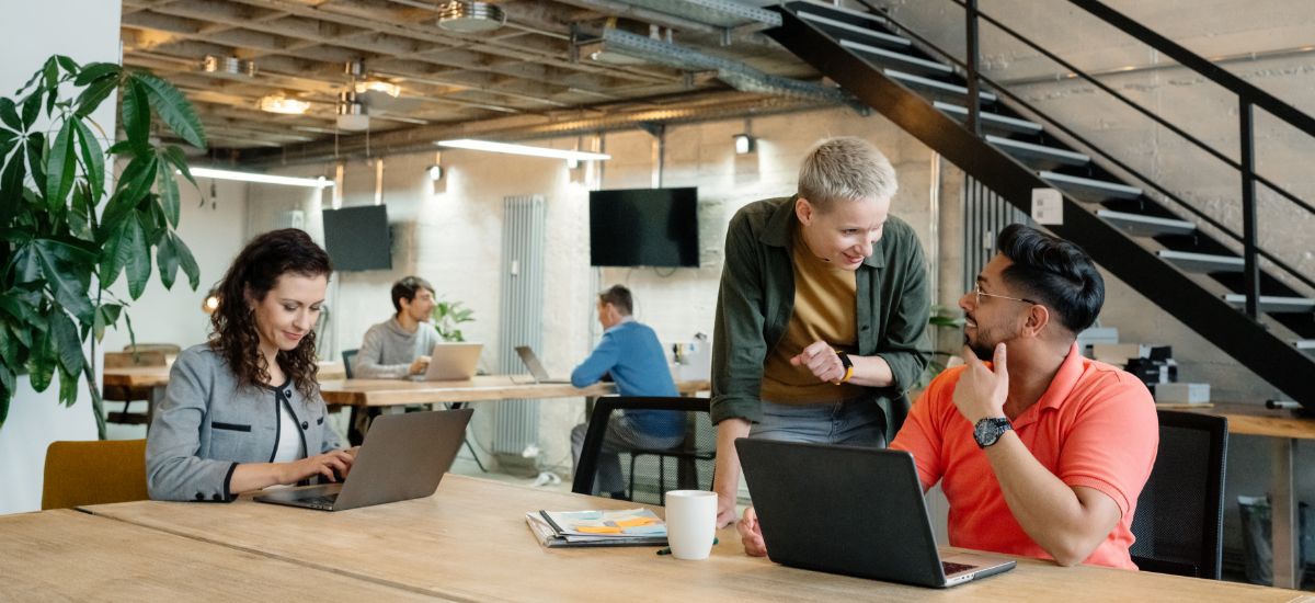 A team of office workers collaborates at a computer, surrounded by colleagues focused on their individual tasks.