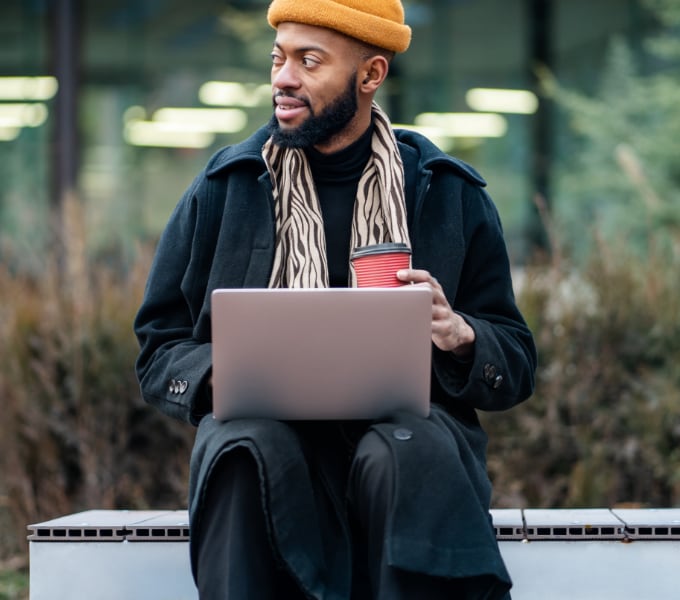 Young African American man working on a laptop computer, sitting on a bench outside and holding a coffee cup.