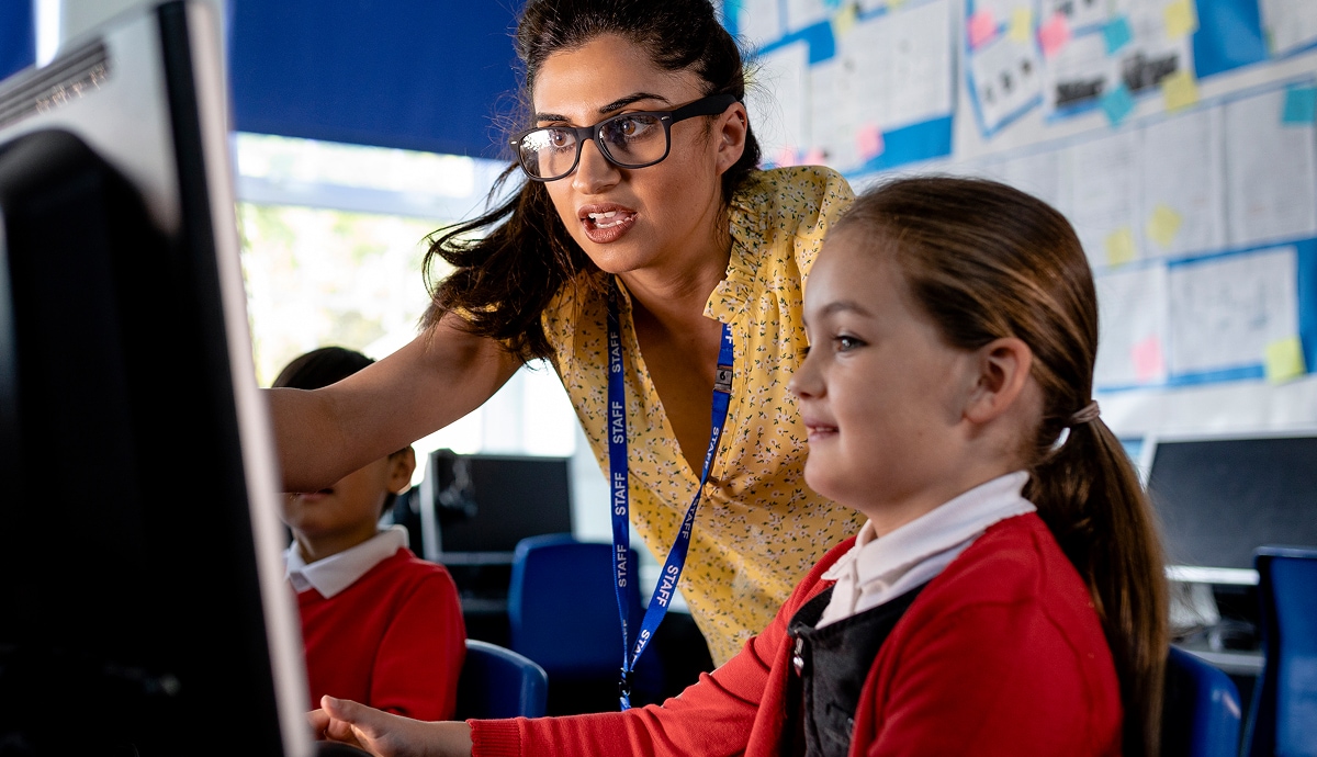 A teacher assists a young student at a computer, pointing at something on the screen.