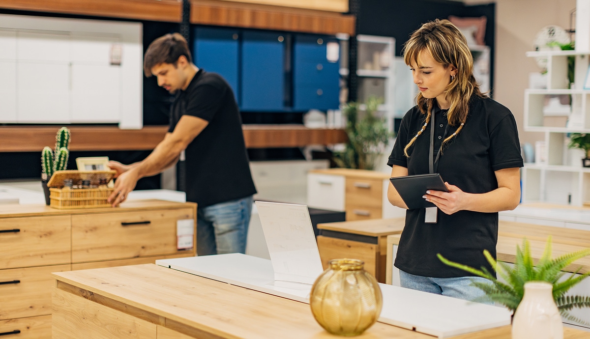 A retail employee uses a tablet while examining a product, as another employee organizes items in the background.