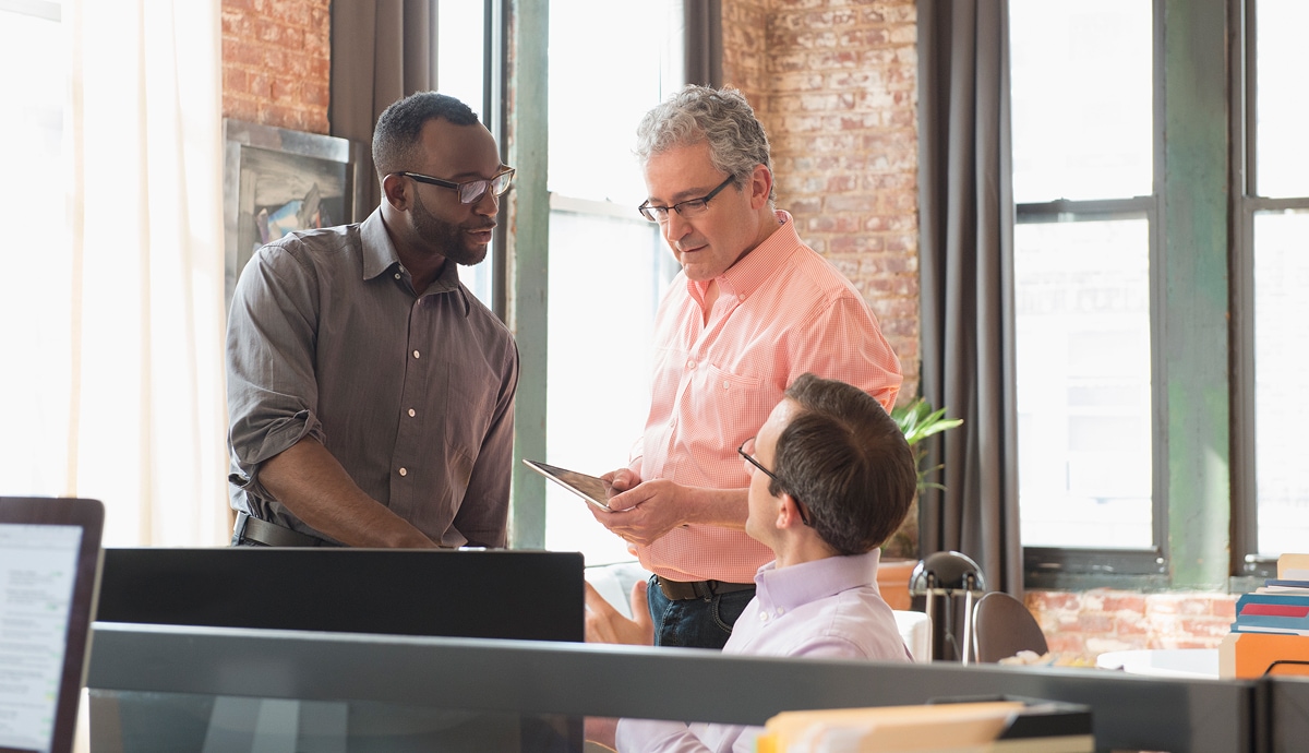Three colleagues discuss a project in a sunny, brick-walled office.