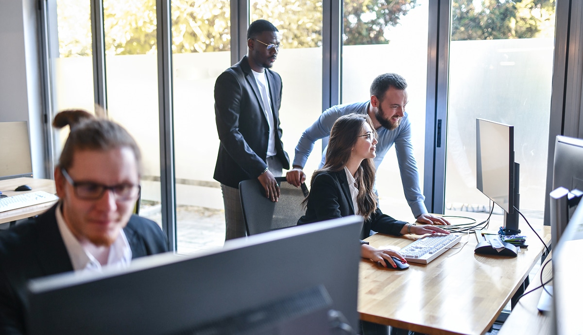 Colleagues collaborate around a computer in a brightly lit modern office.