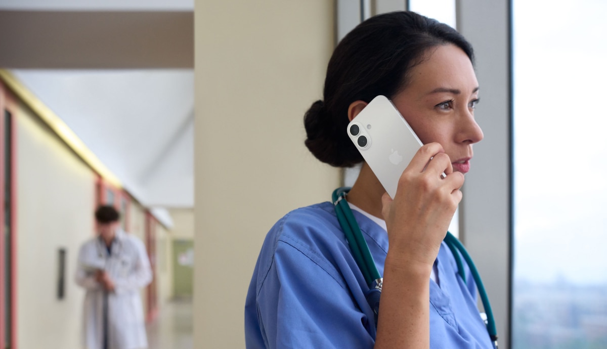 Healthcare professional in scrubs speaks on a smartphone beside a hospital window.