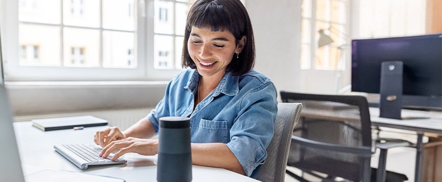 Young businesswoman talking to virtual assistant at her desk. Female professional working on computer and talking into a virtual assistant speaker.