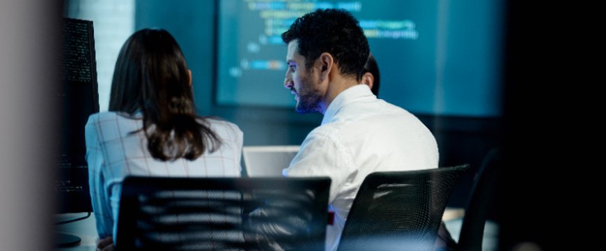 Two professionals seated at monitors reviewing code on large screen in office