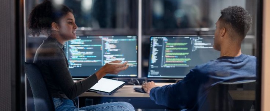 Two professionals collaborating at desk with dual monitors showing code and tablet