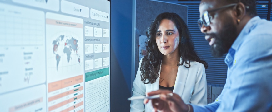 Two office professionals reviewing marketing analytics dashboard displayed on large monitor