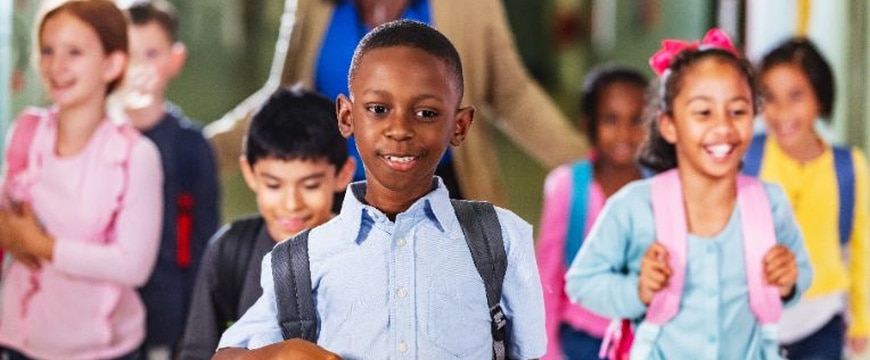 A multiracial group of elementary students walking together with their teacher in the school hallway. The focus is on the African-American boy walking in front.