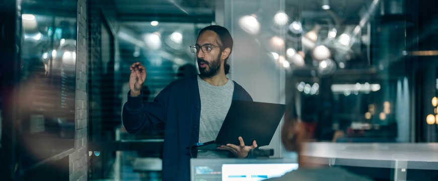 Presenter with laptop speaks near computer workstations in a glass-walled office