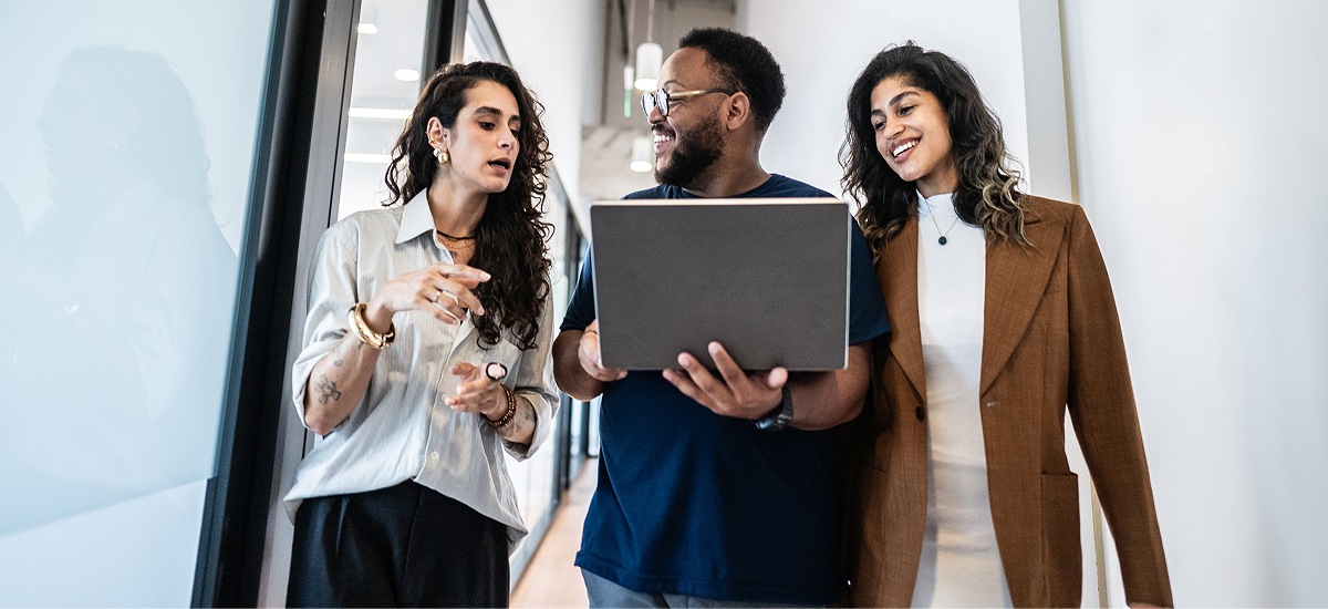 Three colleagues reviewing laptop together in office corridor.