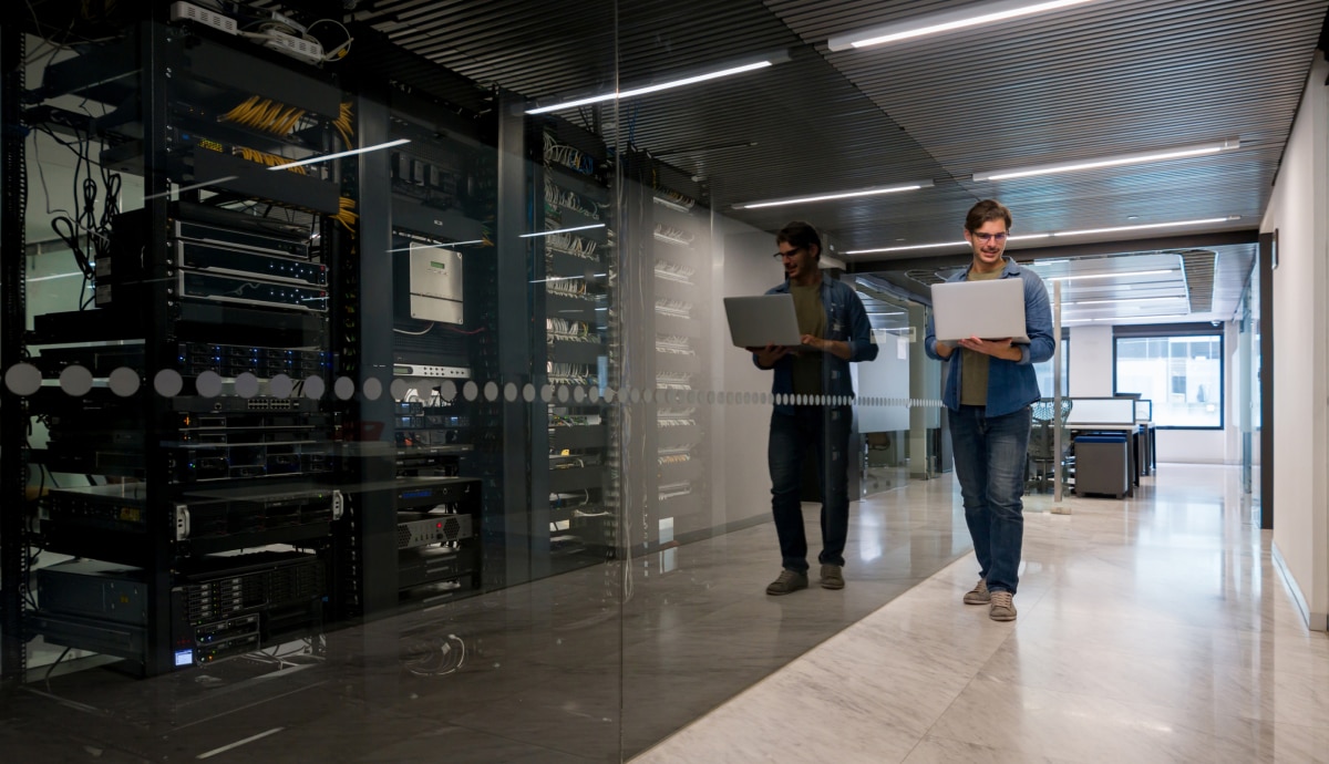IT professionals walk through a data center corridor while monitoring systems on laptops.