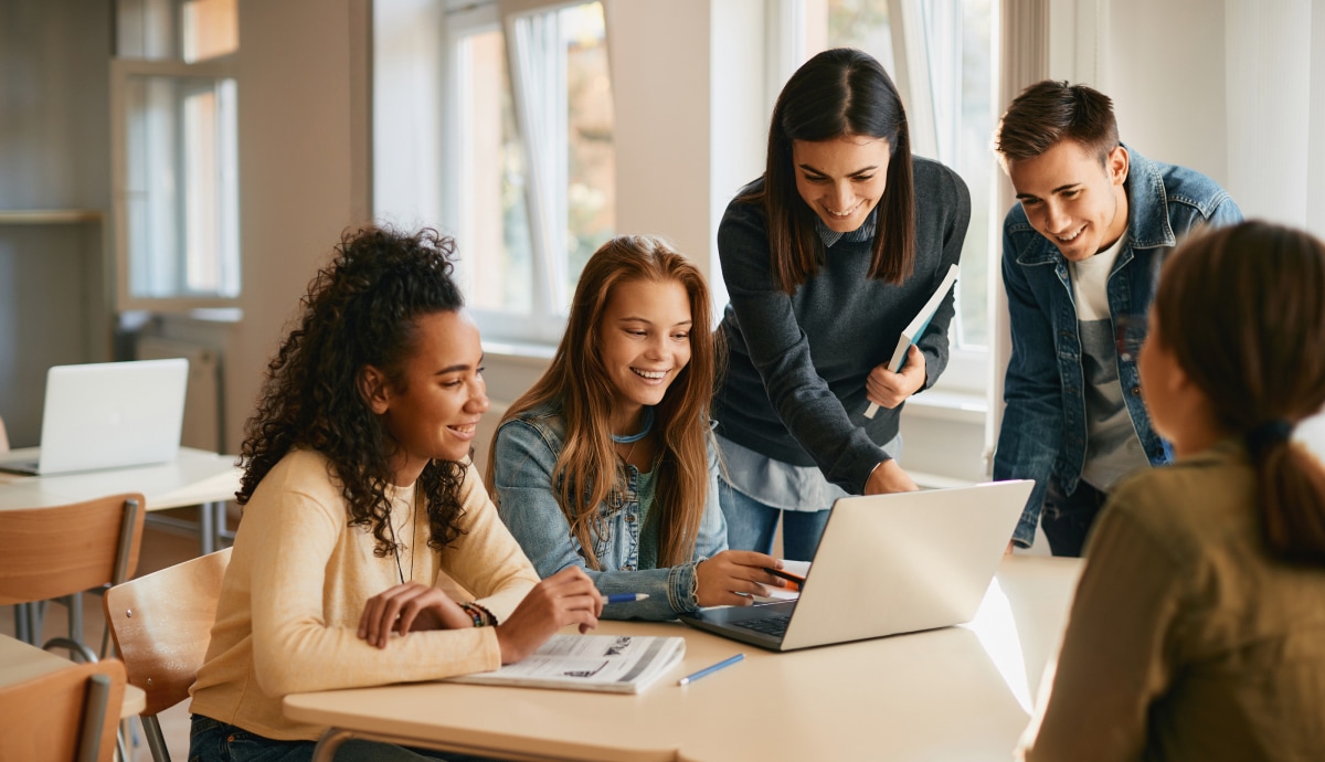 Students collaborate around a laptop during a group activity in a bright classroom.