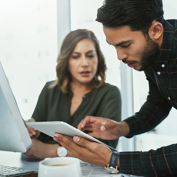 Colleagues analyzing information on tablet beside desktop monitor.