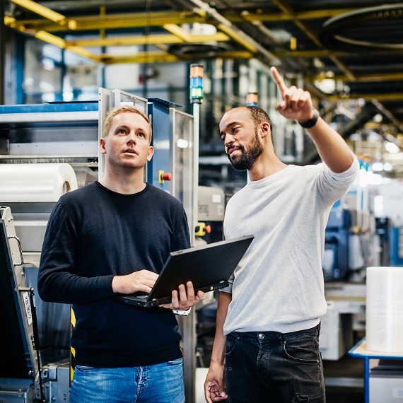 Engineer pointing upward while colleague holds laptop on factory floor.
