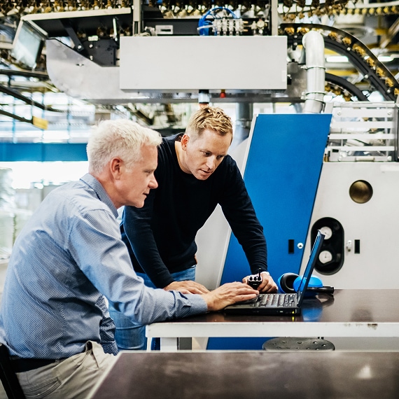 Senior and junior engineers analyzing data at workstation in factory.