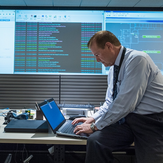 Manager working on laptop with security dashboards displayed behind.