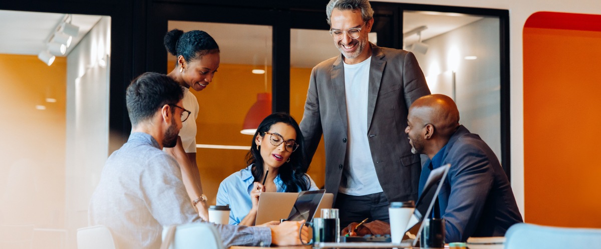 Five colleagues collaborate around a laptop during a workplace discussion.