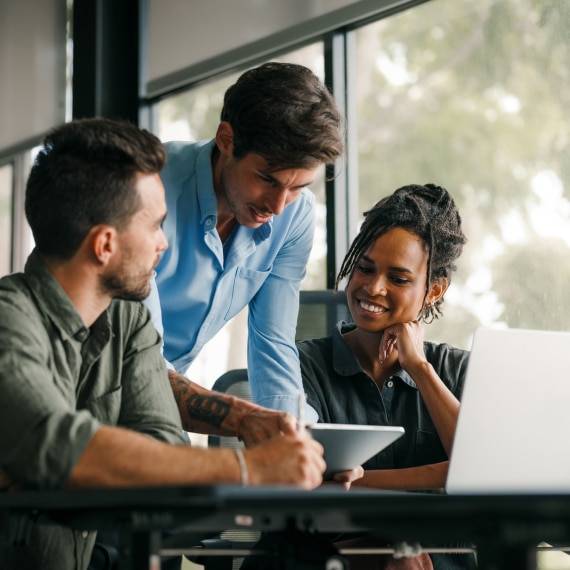 Three colleagues collaborate at a desk, reviewing a tablet and laptop.