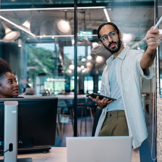 Professional pointing at whiteboard while colleague works at desktop computer.