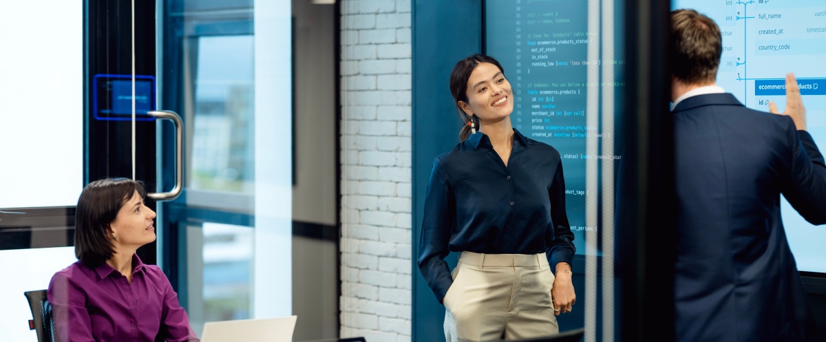 Three colleagues review data and code on a digital board in a modern office meeting room.