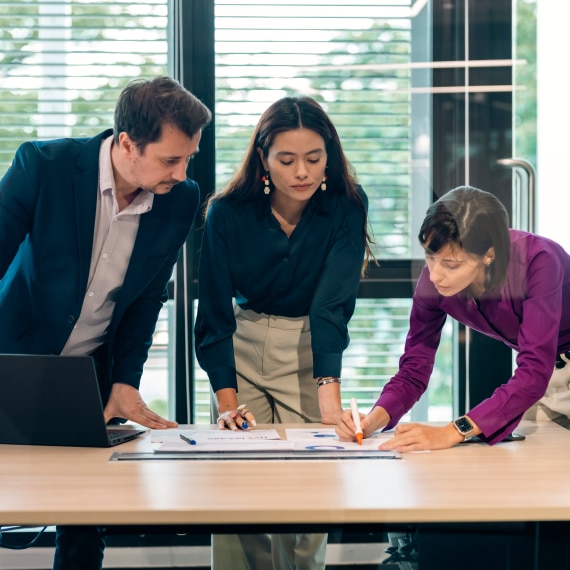 Three colleagues collaborate over printed charts, analyzing data trends in a conference room.