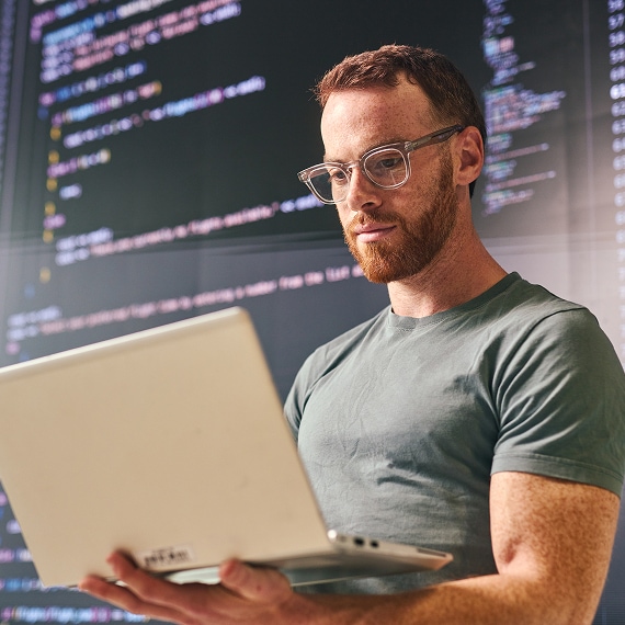 IT professional reviewing code on laptop against large screen display.