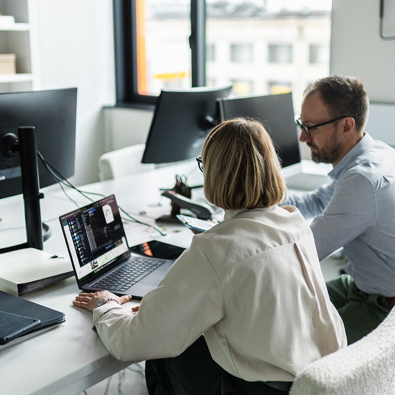Two colleagues collaborating at a desk, reviewing content on a laptop in a bright office.