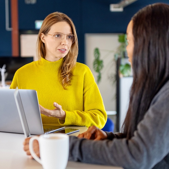 Professional in a yellow sweater discussing work with a colleague across a desk.