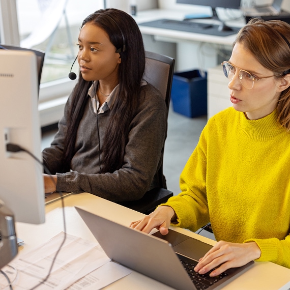 Two support agents working at computers, wearing headsets in a customer service office.