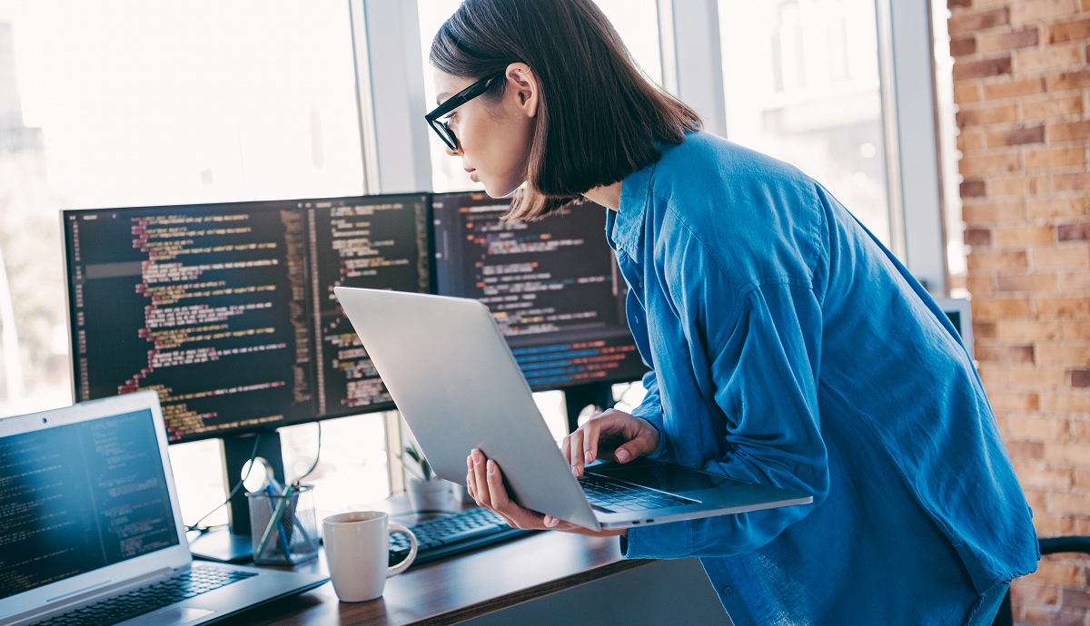 Professional reviewing code on multiple monitors while working on a laptop in a modern office.
