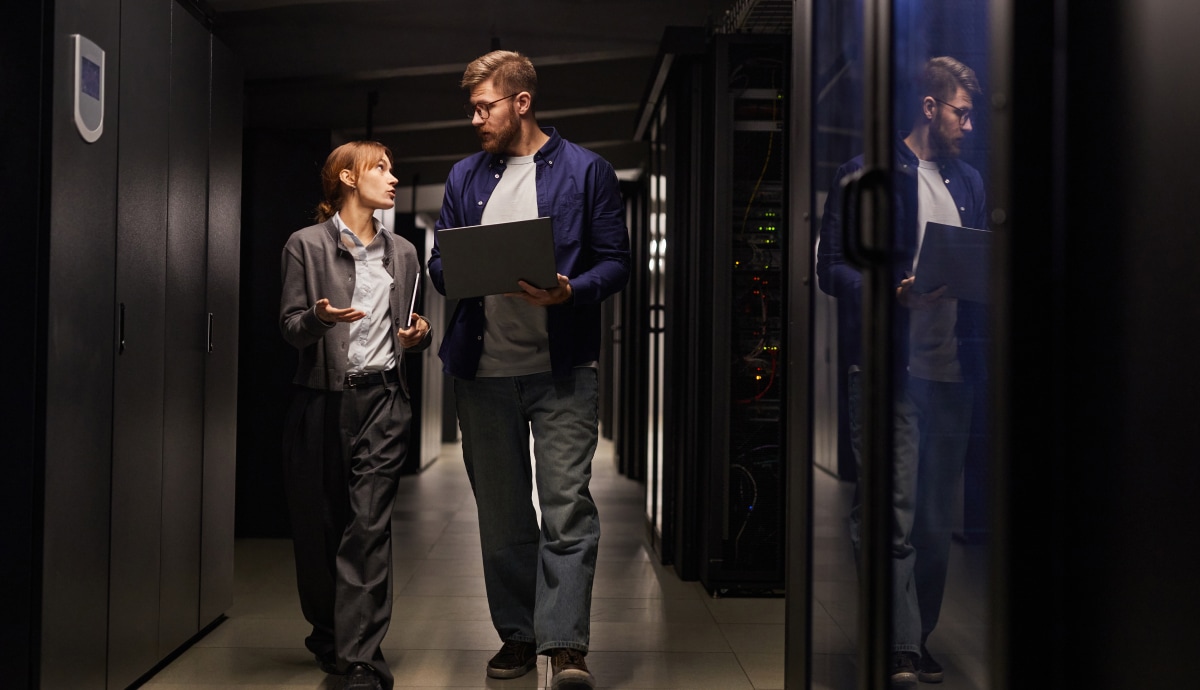 Two IT professionals walk through a data center aisle, reviewing information on a laptop.