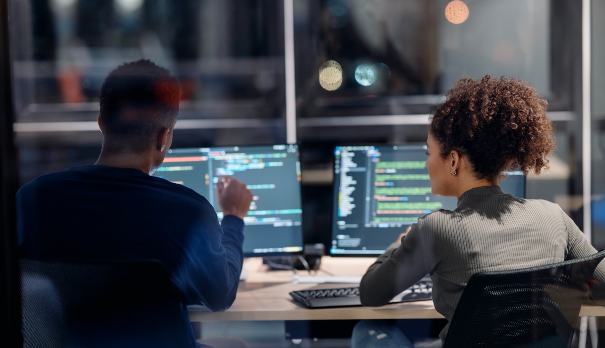 Two IT professionals working together to review and update code on computer monitors.