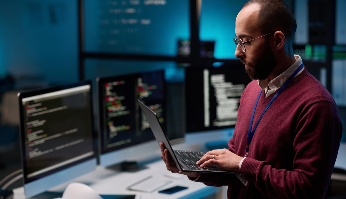 Technology professional works on a laptop surrounded by monitors displaying code.