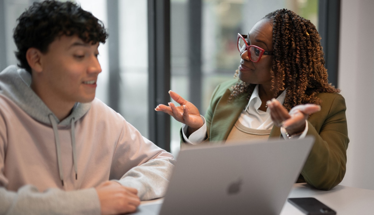 Instructor discusses ideas with student across a laptop in meeting.