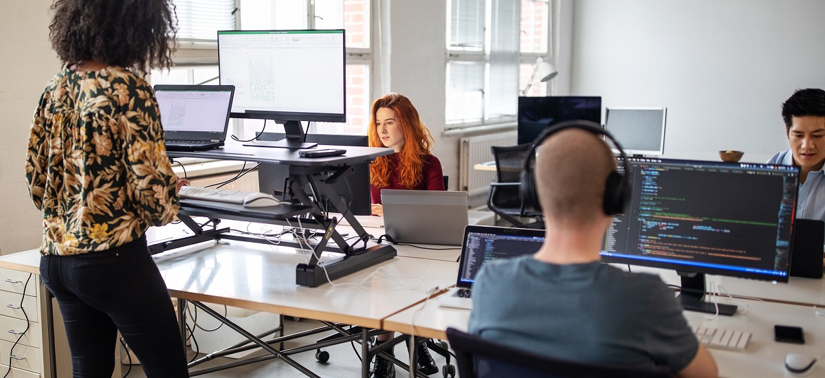 Team collaborating at shared desks with multiple monitors in a bright workspace.