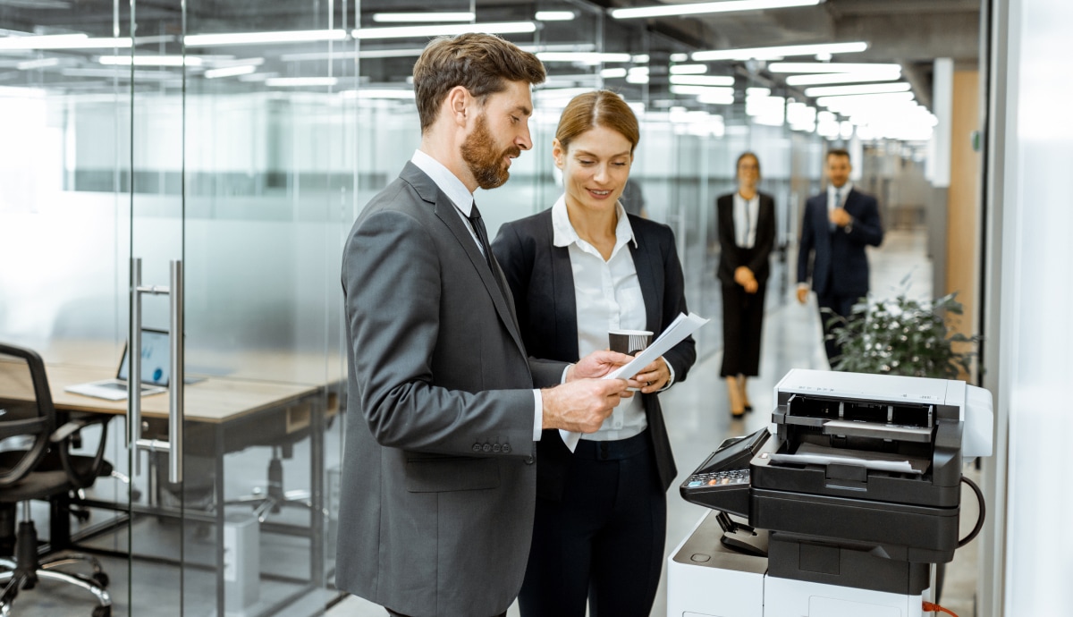 Two colleagues review documents beside an office printer in a modern workplace.