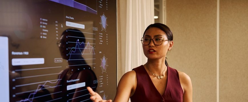 Presenter gestures beside a large screen displaying analytics charts and graphs
