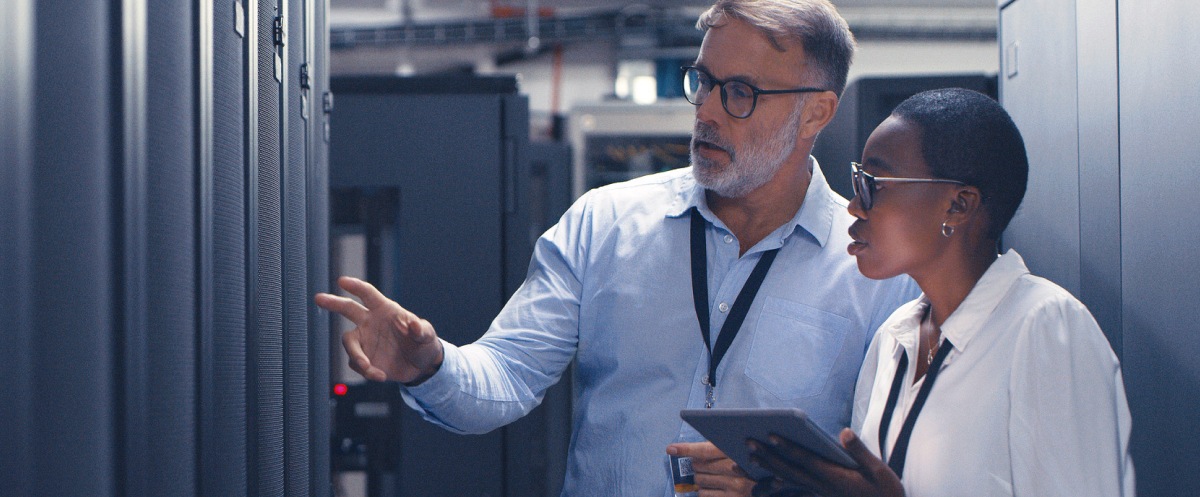 Two IT specialists inspect server racks while reviewing data on a tablet.
